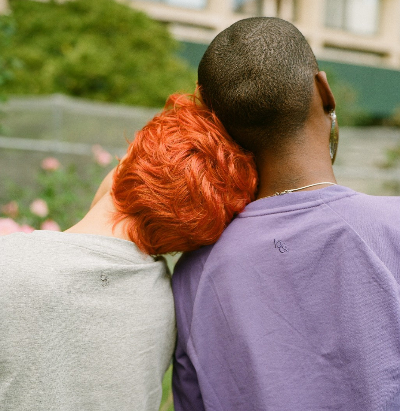 The backs of two people leaning their heads against each other, wearing gray and purple Both& apparel shirts