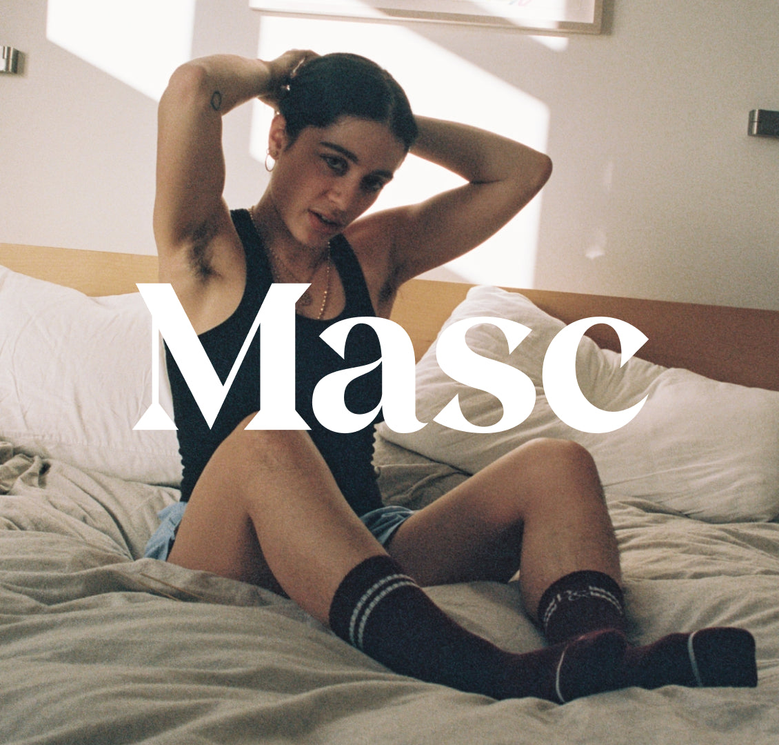'Masc' text over photo of woman wearing black tank, sleep shorts and socks, tying their hair back in bed