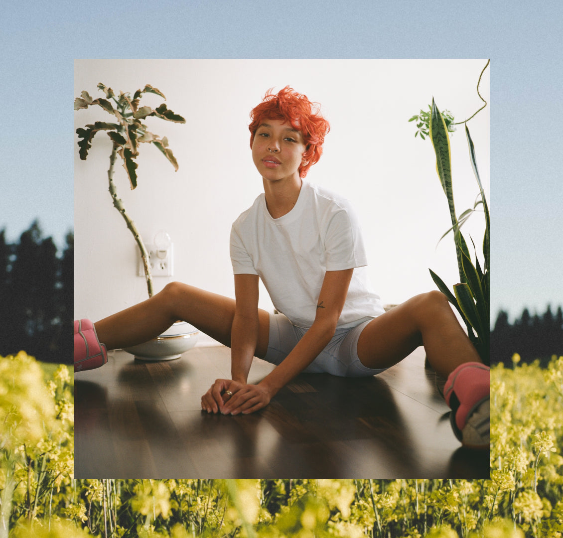 Gender neutral afab person with red orange hair, sitting on floor, surrounded by plants