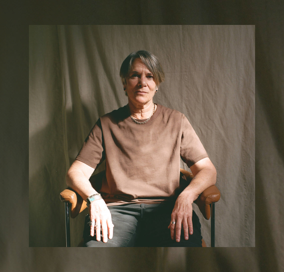 Older queer person in brown shirt sitting in chair against cloth backdrop